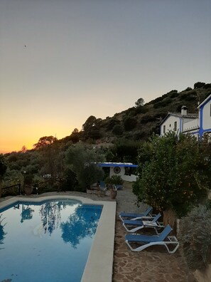 A swimming pool with lounge chairs, surrounded by a hillside with vegetation and a building with blue and white exterior. - Loft type, pool, terrace, wifi, mountain views, sunsets. (Rute)