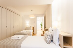 A modern hotel room with a large bed, two smaller beds, a wardrobe, a desk, and a potted plant.