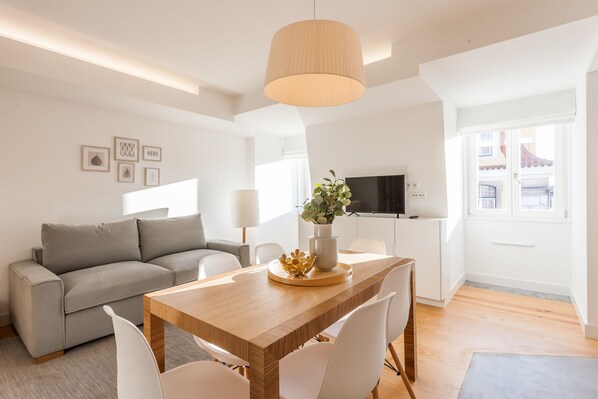 A modern living room with a wooden dining table, white chairs, a grey sofa, and a TV in the background.