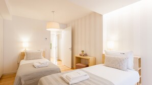 Une chambre d’hôtel avec deux lits, un banc en bois, une petite table et une porte donnant sur une autre pièce.