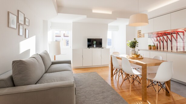 Un salon moderne avec un canapé gris, une table à manger en bois avec des chaises blanches, un téléviseur posé sur un meuble blanc et un coin cuisine avec un plan de travail rouge.