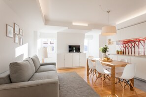 Een moderne woonkamer met een grijze bank, een houten eettafel met witte stoelen, een televisie op een wit kastje, en een keuken met een rode achterwand.