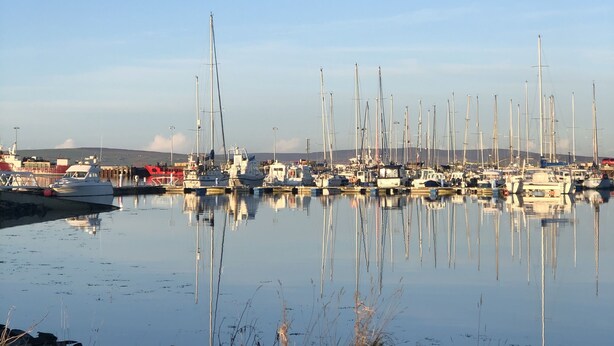 A marina with multiple boats docked, calm water, and rocky shoreline.