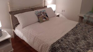 A neatly made bed with a white comforter and two pillows, a bedside table with a lamp, and a wall-mounted power outlet.