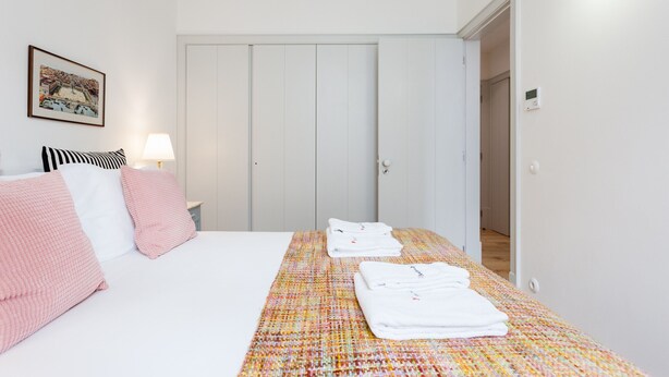 A hotel room with a bed, pillows, a bedside table with a lamp, a wall-mounted picture, and a door. - FLH Bacalhoeiros Charming Place (Lisbon)