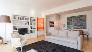 A modern living room with a white sofa, a black rug, a bookshelf, a flat-screen TV, and a large abstract painting. - FLH Chiado Design Flat (Lisbon)