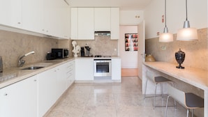 A modern kitchen with white cabinets, a built-in oven, a microwave, and a sink. There are two bar stools and pendant lights above the counter. - FLH Chiado Design Flat (Lisbon)
