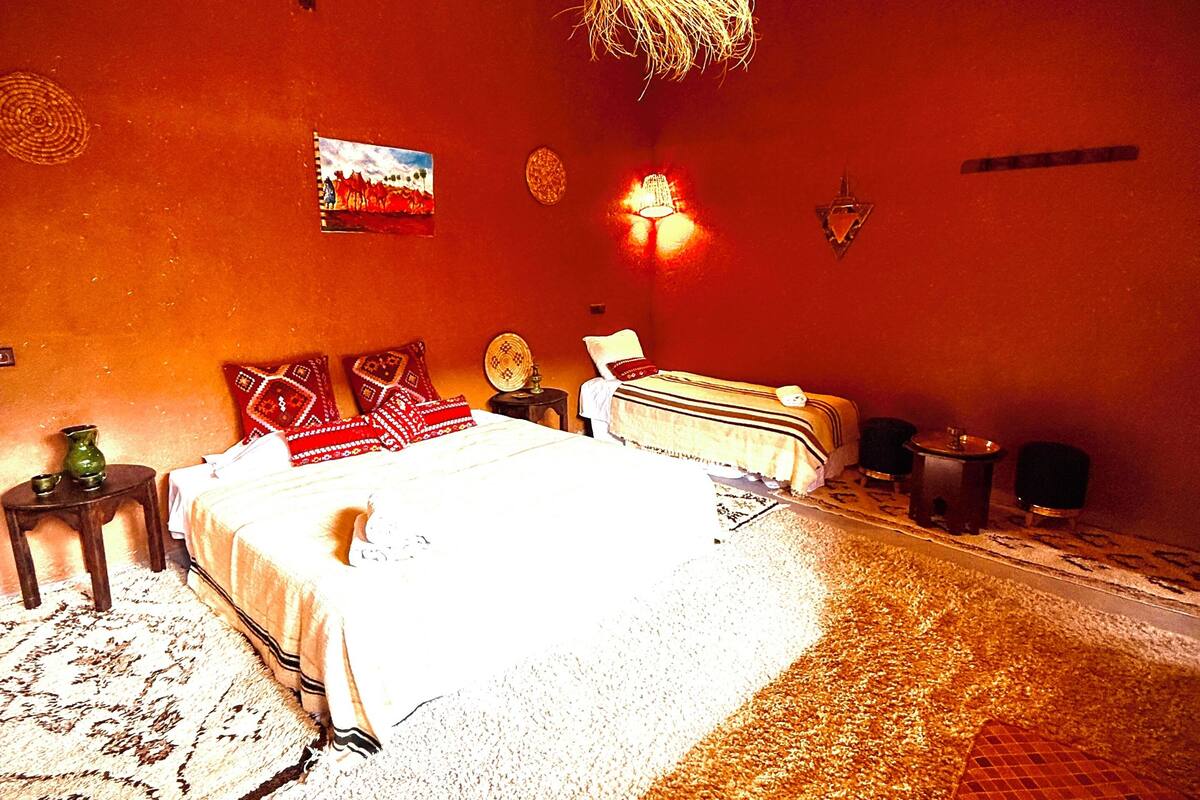 Um quarto com uma cama, uma mesa pequena, uma luminária na parede e um lustre decorativo.