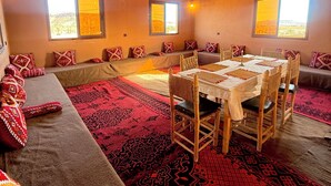 A cozy room with a wooden ceiling, a dining table with chairs, a sofa with patterned cushions, and two windows with a view of the landscape. - Dar Talmoudi (Tagounite)