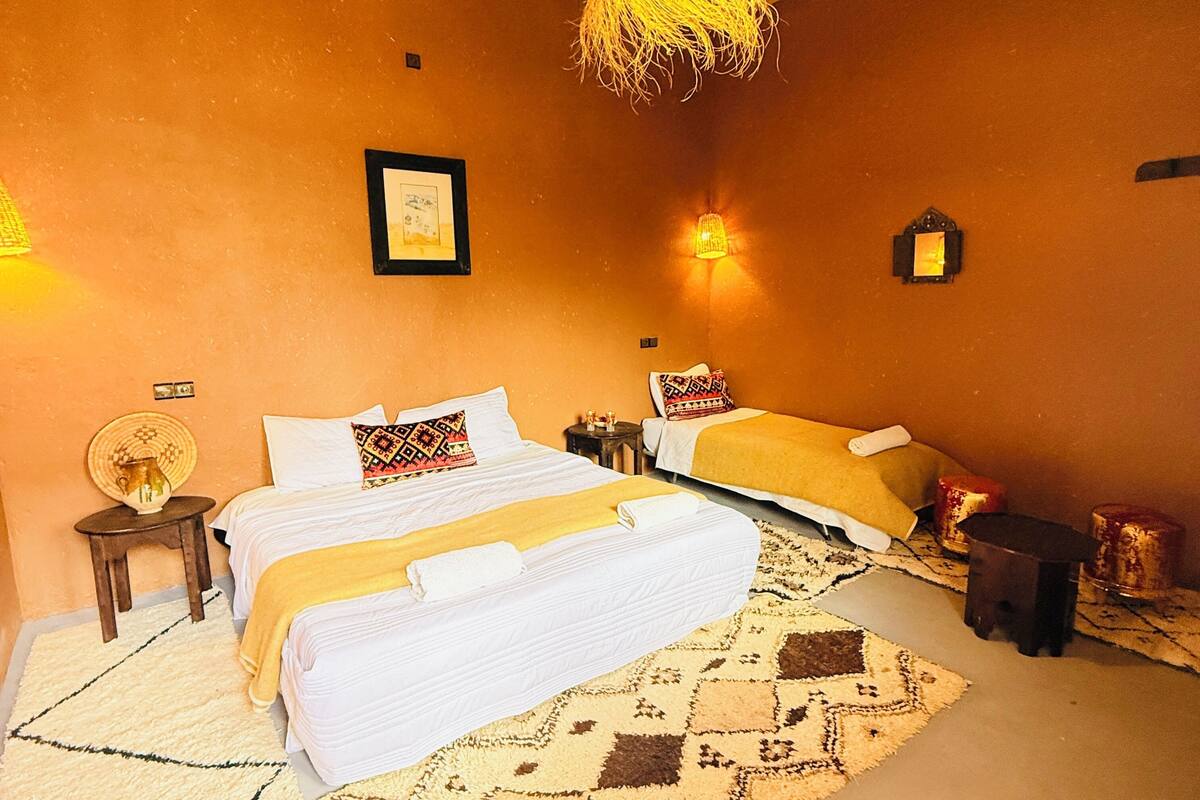 Quarto com teto de tecido, duas camas com roupa branca, tapete estampado e luminárias fixadas na parede.