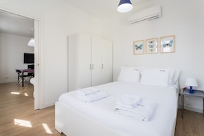 A clean, modern bedroom with a large bed, white linens, a wardrobe, a desk with a chair, and a flat-screen TV.