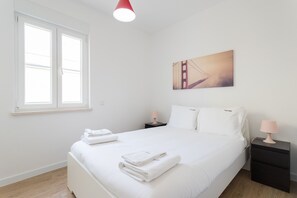 Ein ordentlich bezogenes Bett mit gefalteten Handtüchern, ein Nachttisch mit Lampe, ein Fenster mit Jalousien und ein Bild einer Brücke an der Wand.
