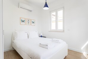 A clean, white hotel room with a bed, a window, and wall-mounted air conditioning.