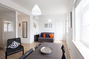 Ein modernes Wohnzimmer mit Esstisch, einer grauen Couch mit orangefarbenen Kissen und einem schwarzen Sessel mit gemustertem Kissen.