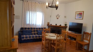 Uma sala de estar com sofá, mesa de jantar, cadeiras, lareira, televisão e lustre.