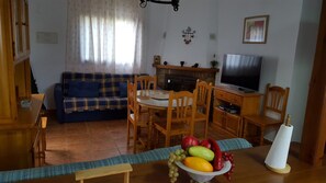 Uma sala de estar com sofá, mesa de jantar com cadeiras, televisão e uma cesta de frutas.