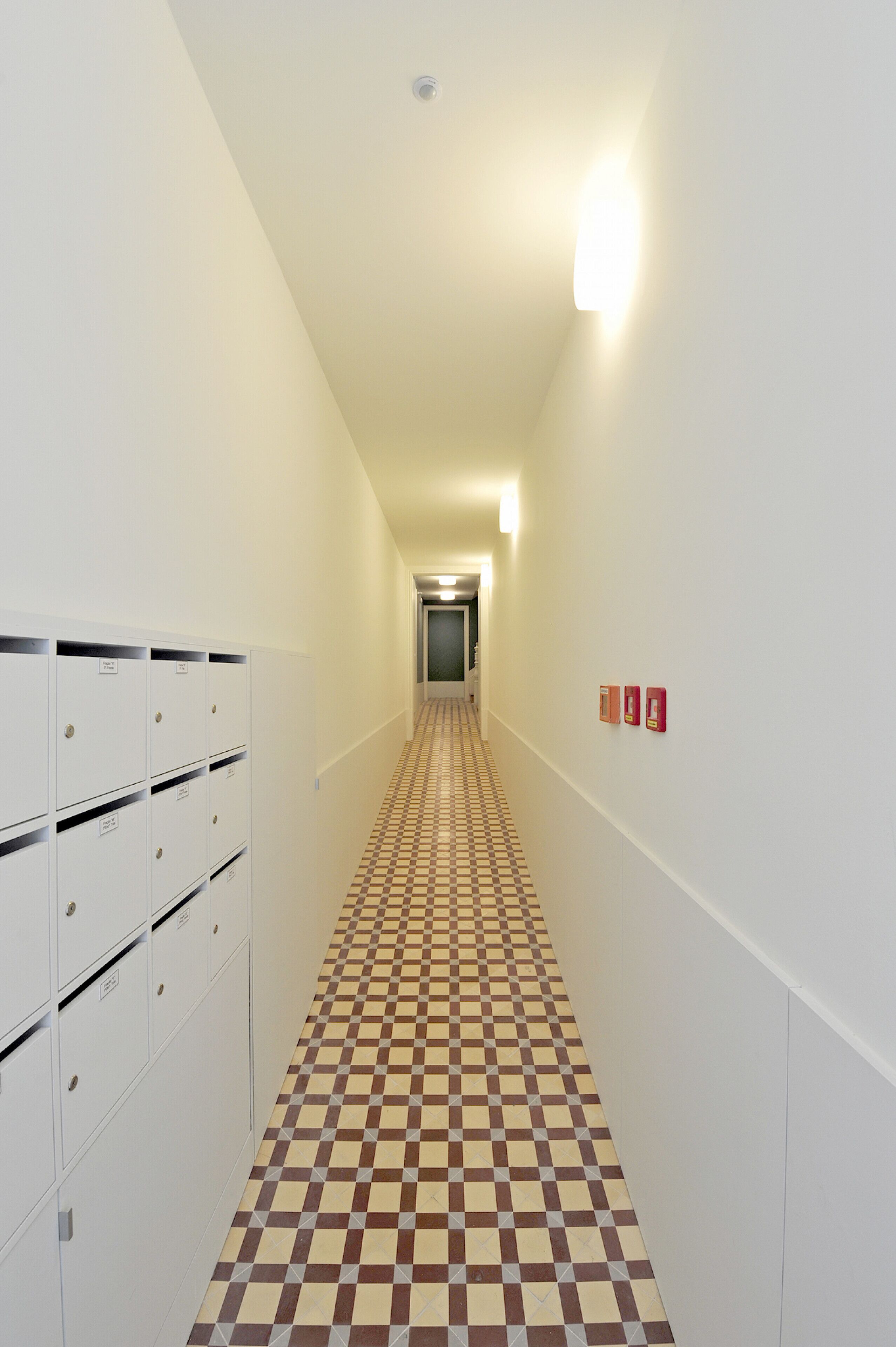 Un couloir avec des casiers, un sol à carreaux et des panneaux indiquant les sorties de secours.