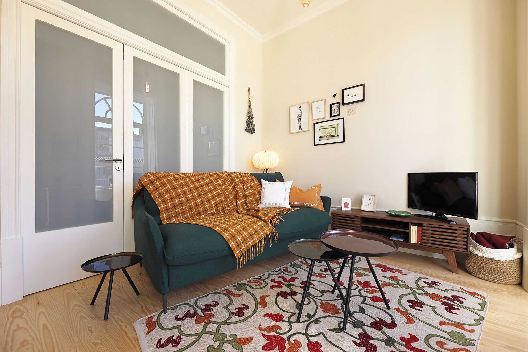 Un salon avec un canapé vert, un tapis à motifs, une petite table d’appoint, un téléviseur et une étagère murale décorée de tableaux.