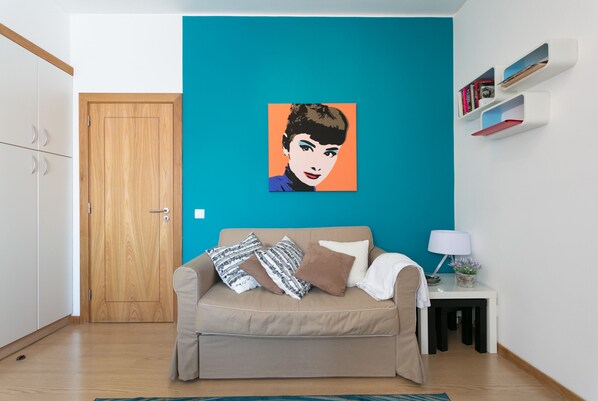 A living room with a beige sofa, a teal accent wall, a wooden door, and a small white side table with a lamp and books.