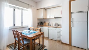 Una cocina compacta con muebles blancos, un refrigerador, una cafetera y una mesa de comedor con sillas.