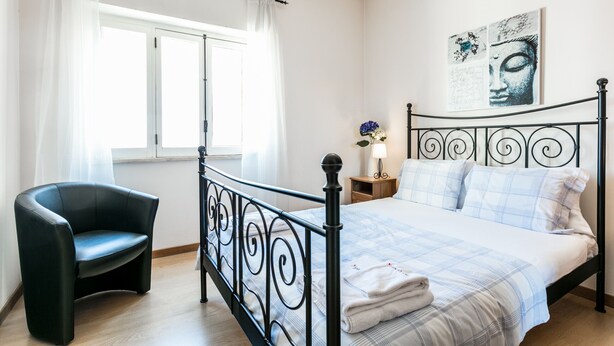 Un dormitorio con cama, una silla, una mesita de noche y una ventana con cortinas.