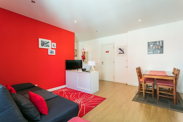 A modern living room with a red accent wall, a black sofa with red cushions, a white entertainment unit, and a dining area with a wooden table and chairs.