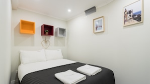 Habitación de hotel con cama, dos almohadas, una colcha negra, dos toallas dobladas, un estante pequeño con una planta, un marco de foto y un estante en la pared.