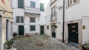 Un estrecho patio empedrado con edificios a ambos lados, destacando puertas y contraventanas verdes, y una pequeña planta en maceta.