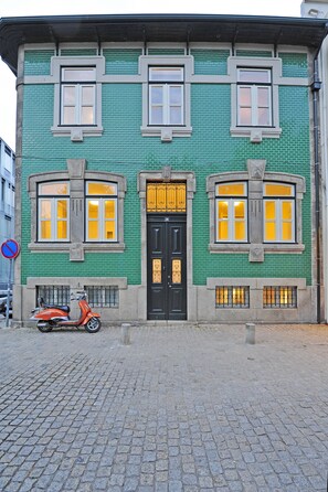 A green brick building with a black door, three windows with yellow frames, and a scooter parked in front.
