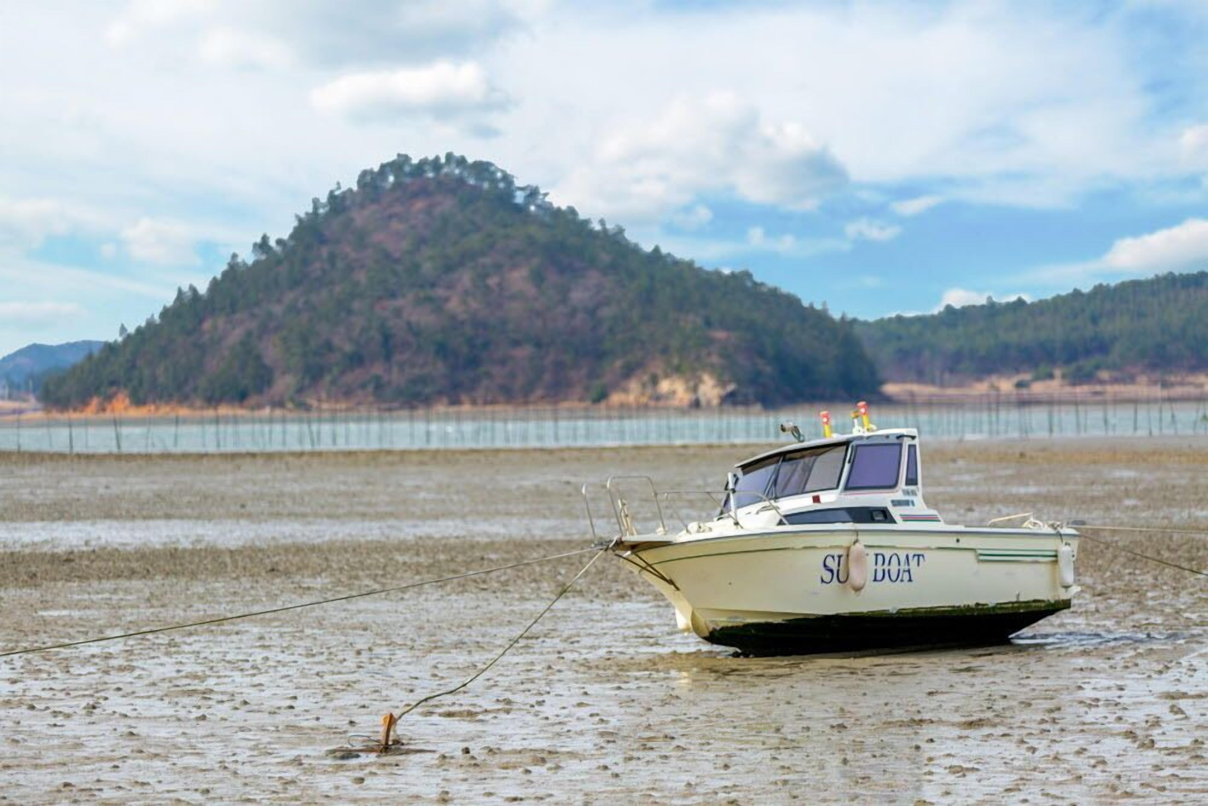 Un bateau baptisé « SUN BOAT » était amarré sur une rive boueuse, avec une colline boisée en arrière-plan.