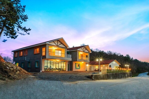 A modern two-story building with large windows and a balcony, surrounded by a natural landscape.