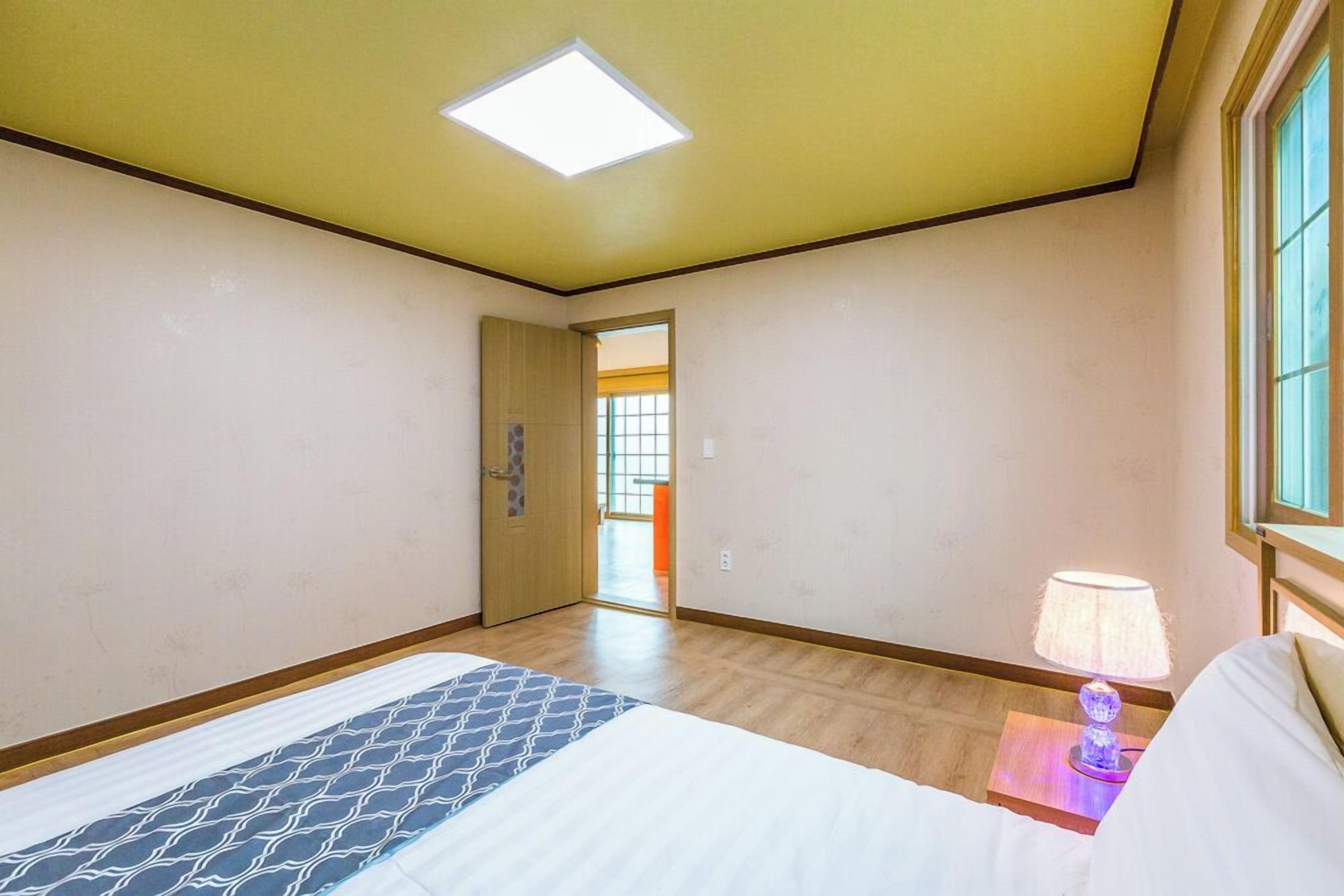 A hotel room with a bed, a nightstand, a lamp, a ceiling light, and a door leading to another room.