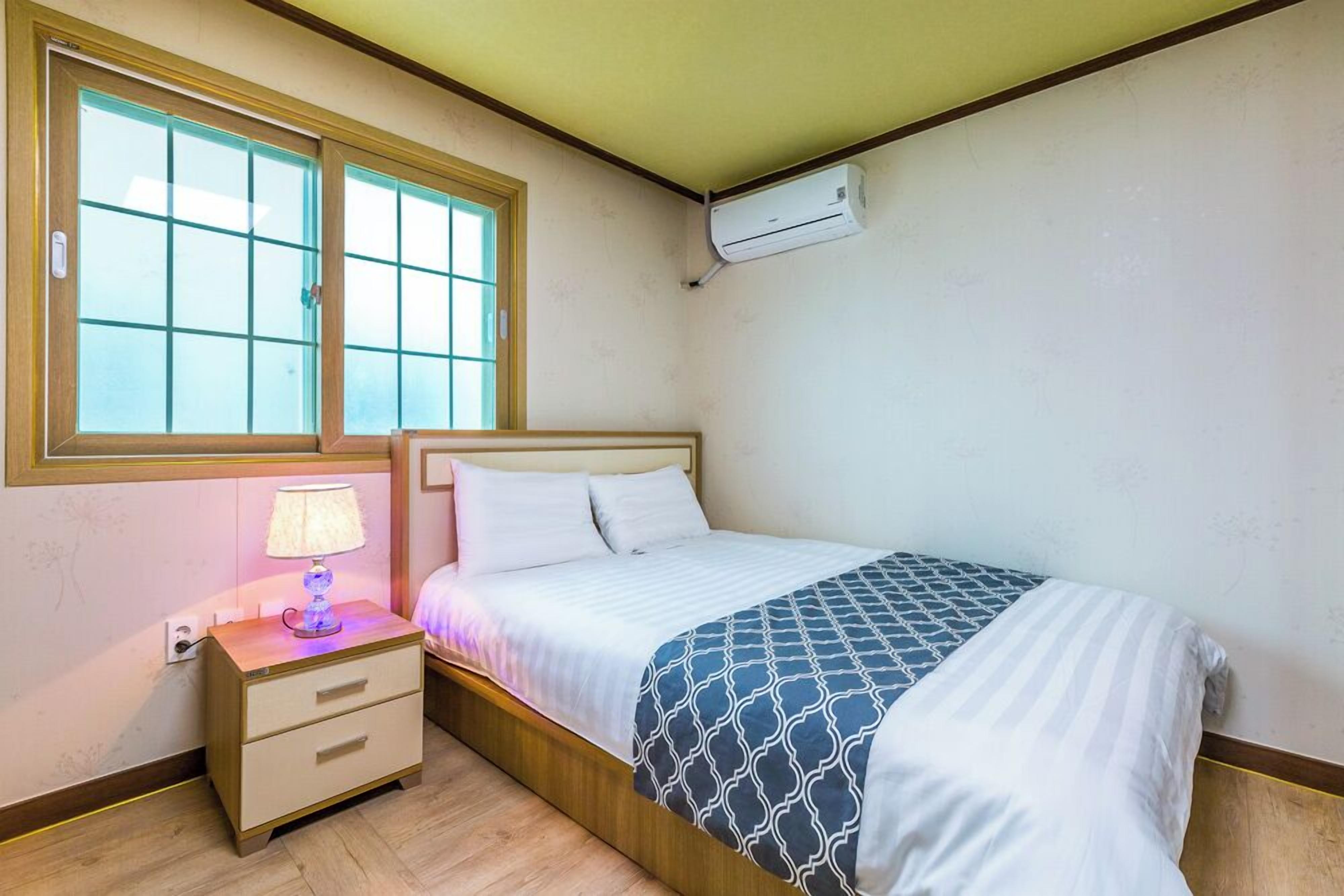 Ein Schlafzimmer mit Bett, Nachttisch, Lampe, Fenster und Klimaanlage.