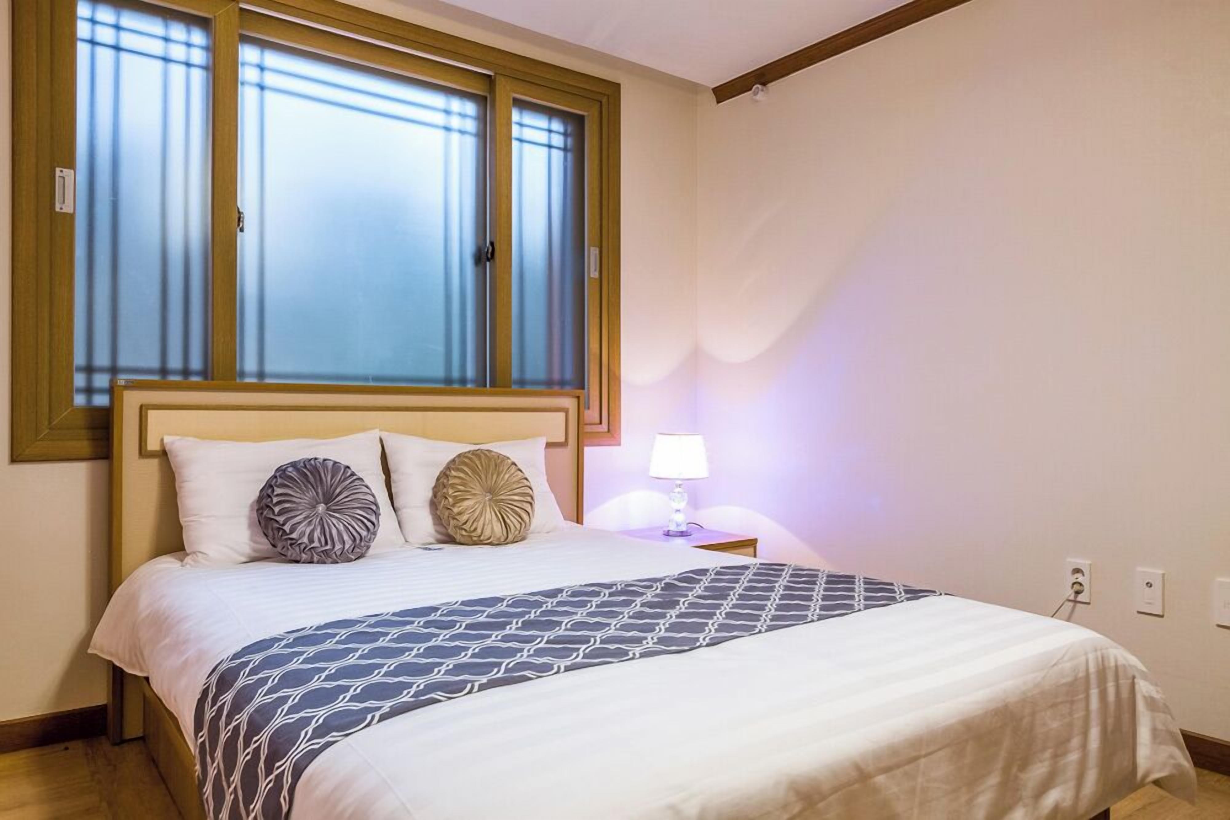 A bedroom with a bed, a nightstand, a lamp, and a window with blinds.