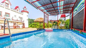 Una piscina con zona cubierta, un edificio parecido a un castillo y una construcción con techo de paja al fondo.
