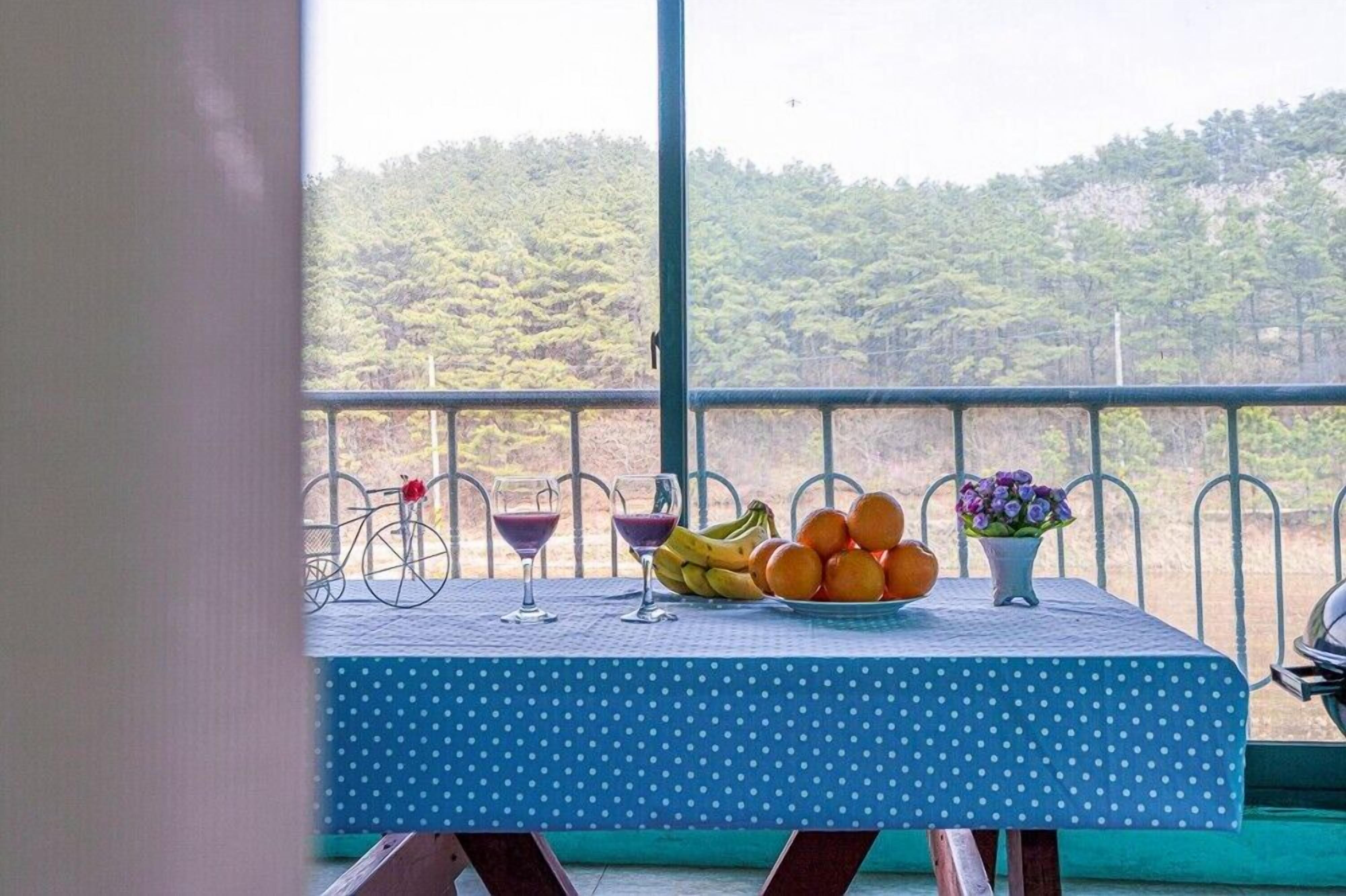Un balcón con una mesa preparada para dos personas, con un mantel de lunares azules, un plato de fruta, dos vasos y un jarrón con flores.