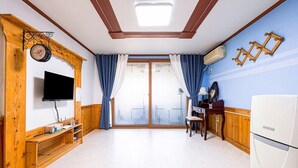 Una habitación con televisión, muebles de madera, refrigerador y un ventanal grande con cortinas.