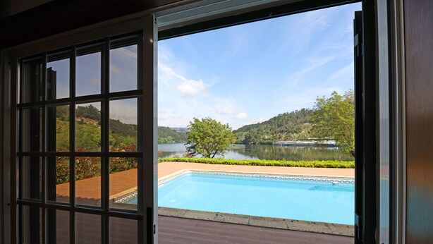 A view from a room with a large window overlooking a swimming pool, a lake, and a hilly landscape.