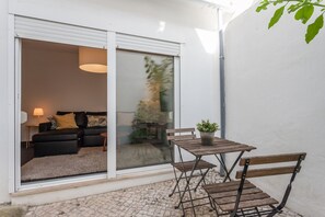 Un patio con una mesa de madera y bancos, una planta en maceta y un ventanal corredero que da a una sala interior con un sofá y una lámpara.