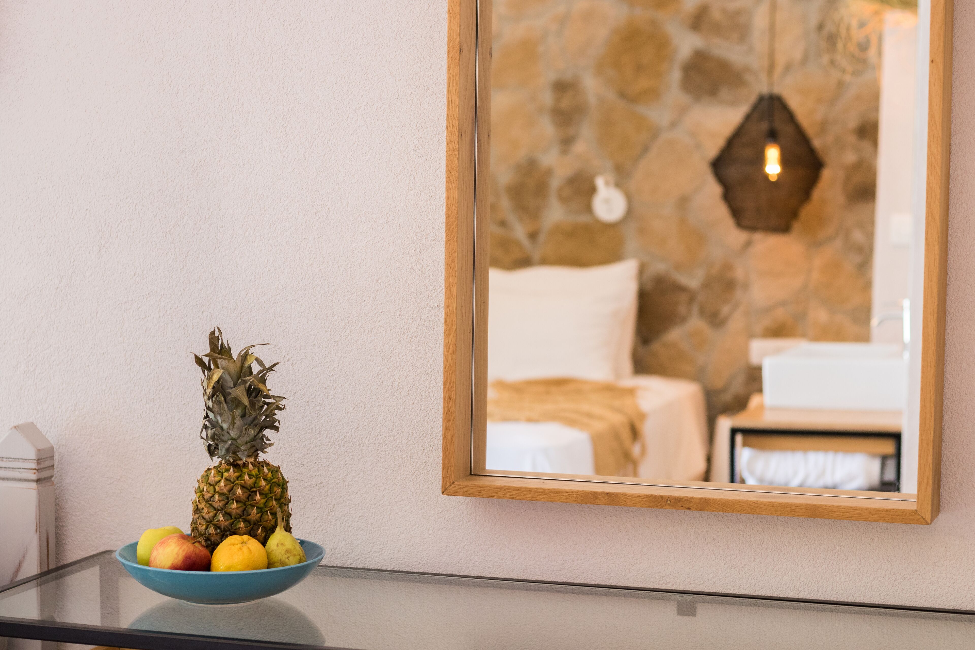 Una ciotola di frutta con ananas e mele su un tavolo di vetro, uno specchio con cornice in legno che riflette un muro in pietra e un letto.