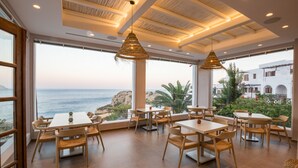 Een restaurant met tafels en stoelen, grote ramen met uitzicht op zee en een rotsachtig landschap.