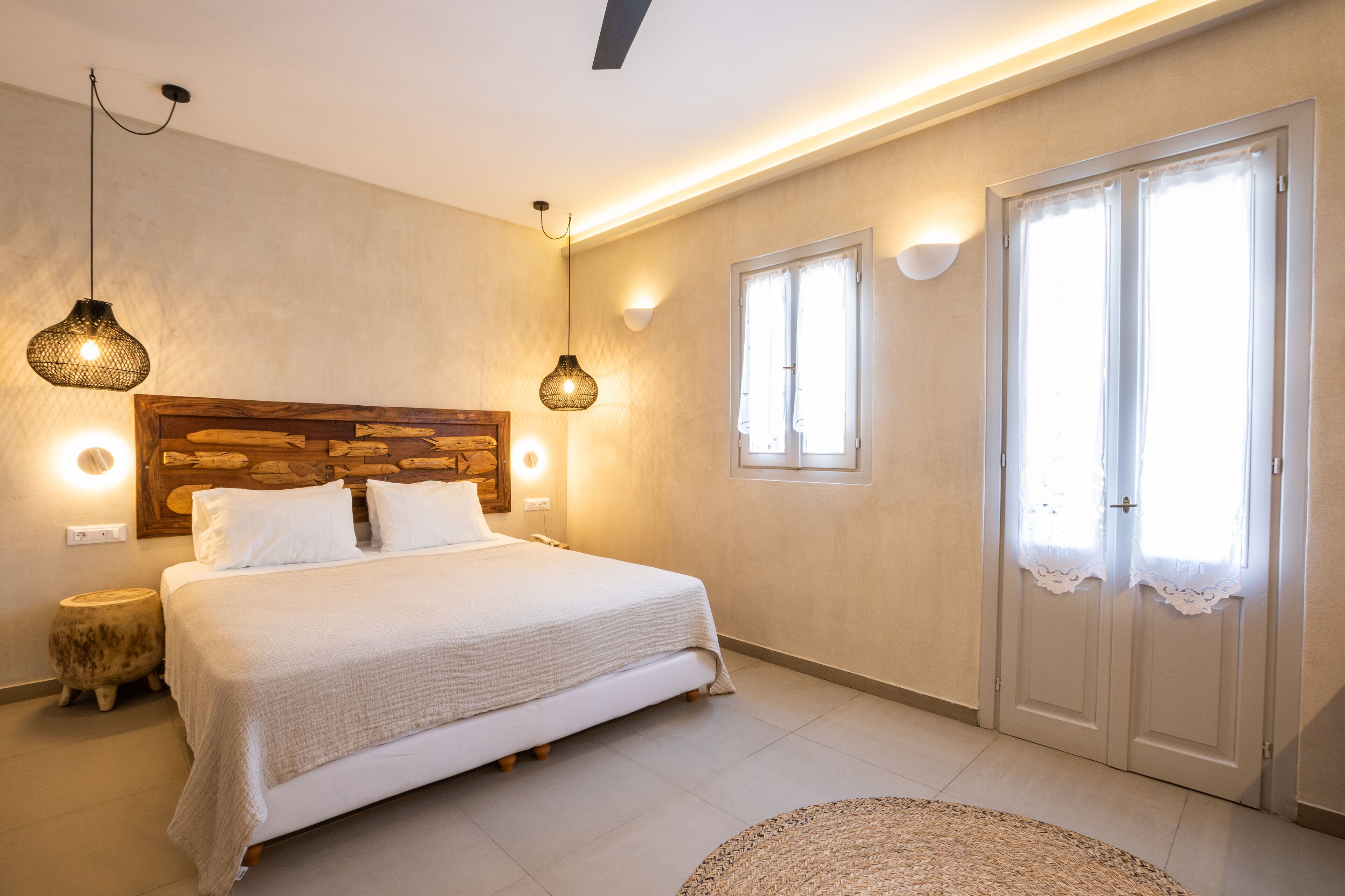 Une chambre avec une tête de lit en bois, un lit avec une literie blanche, deux lampes suspendues, un tapis rond et une porte double avec des rideaux.