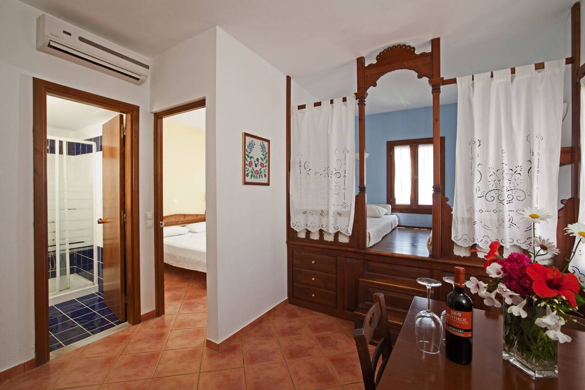 A hotel room with a bed, a wooden dresser, a mirror, a table with a wine bottle and flowers, and a bathroom with a shower and a blue-tiled floor.