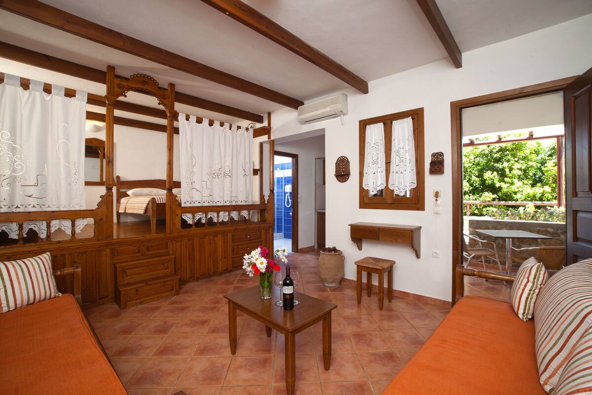 A cozy room with a wooden bed, a small table with a bottle and flowers, and a view of greenery outside.