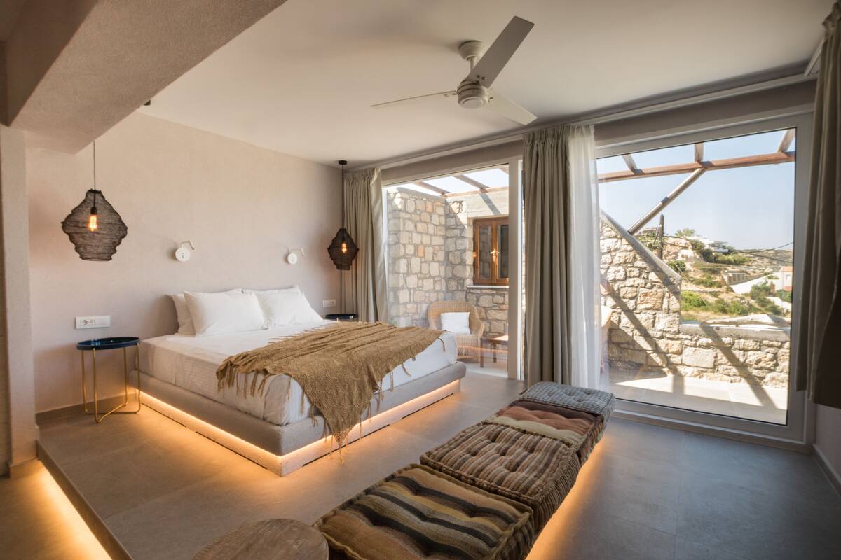 A modern bedroom with a large bed, a bench, and a view of a stone building through a glass door.
