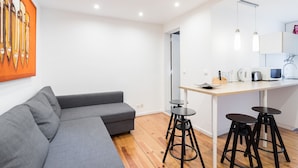 A modern living space with a grey sofa, a wooden dining table with stools, and a kitchen area with appliances. - FLH Lx Factory Modern Flat (Lisbon)