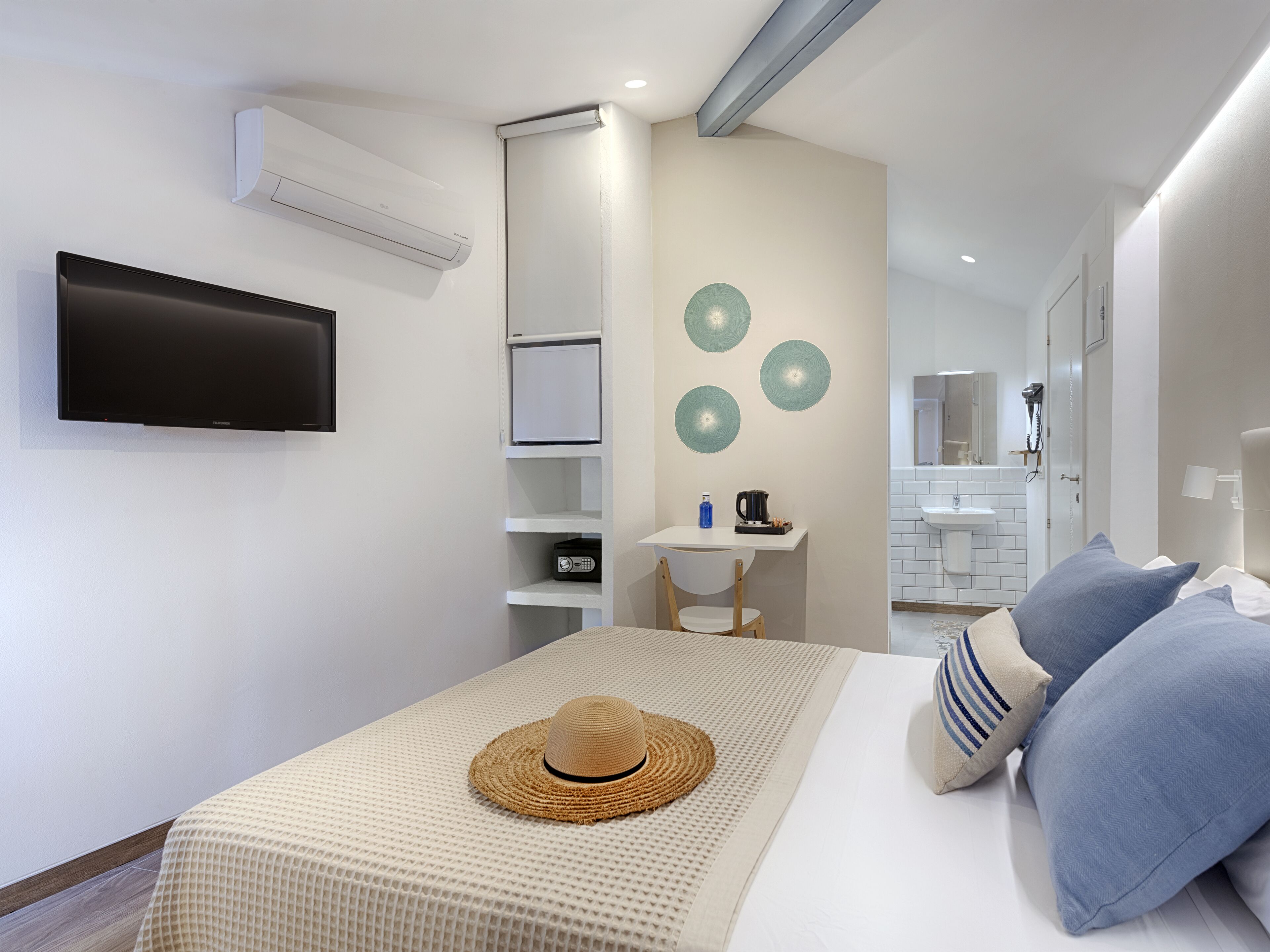 Una habitación de hotel moderna con una televisión de pantalla plana, un escritorio pequeño con una silla, una cama con almohadas azules y a rayas, y un aire acondicionado instalado en la pared.