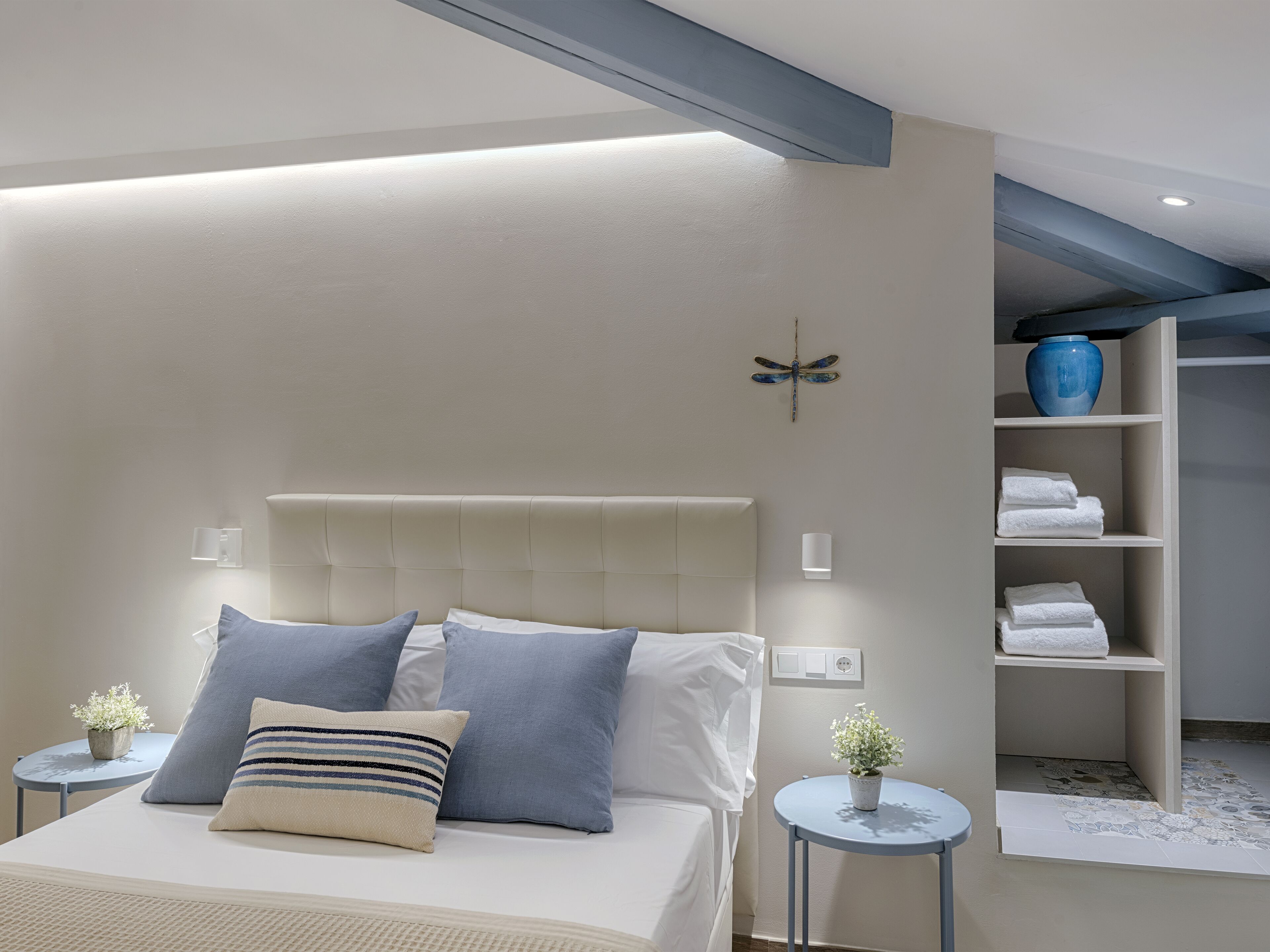 A modern bedroom with a tufted headboard, a bed with white linens, blue and striped pillows, bedside tables with plants, and a blue vase on a shelf.