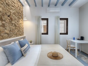 A bedroom with a stone wall, a bed with blue and striped pillows, a woven hat, a small table with a blue bottle and a black coffee maker, and two windows with curtains.