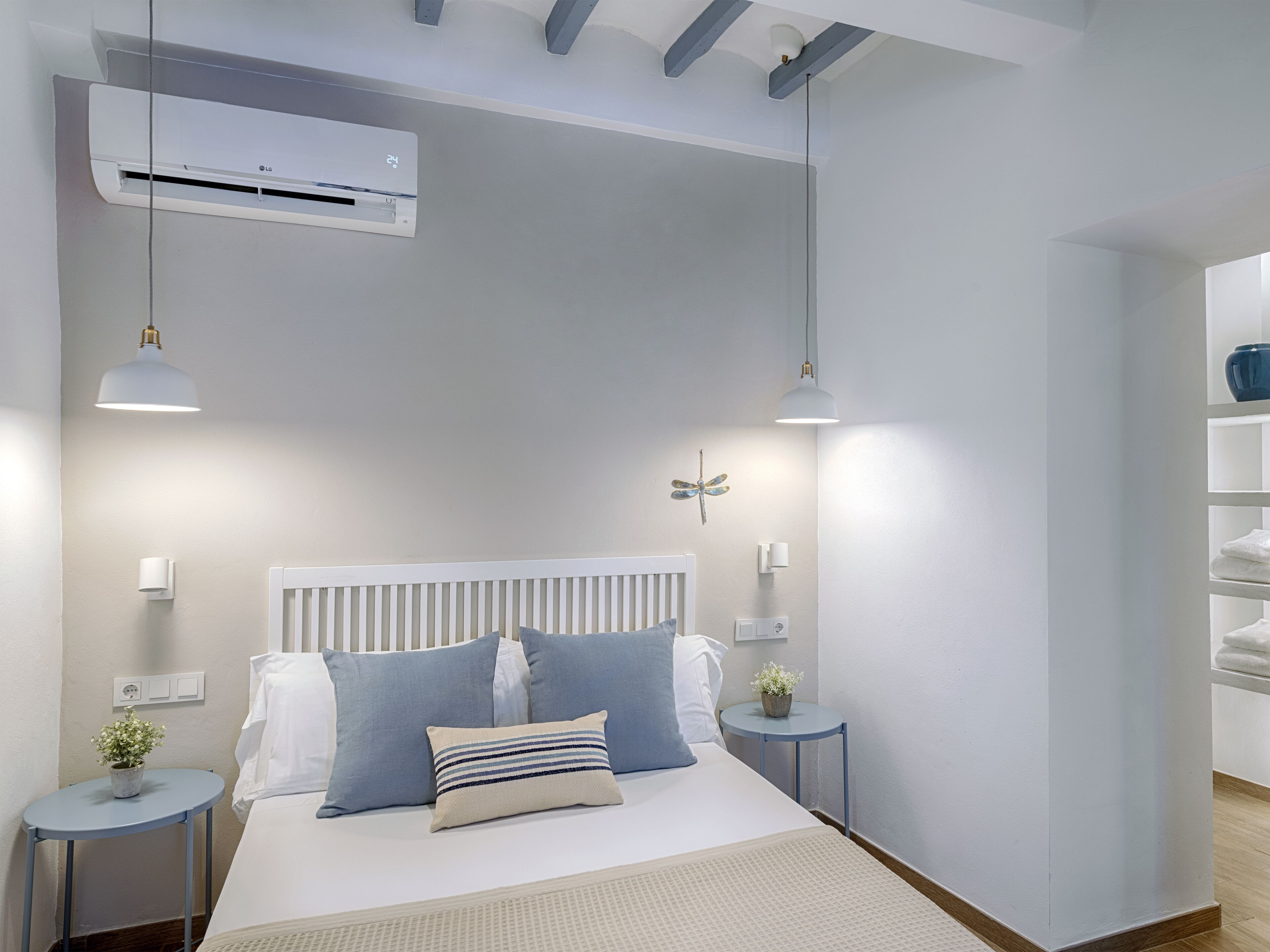 Quarto moderno com cama branca, almofadas azuis e brancas, uma mesa redonda pequena com uma planta e um radiador branco.
